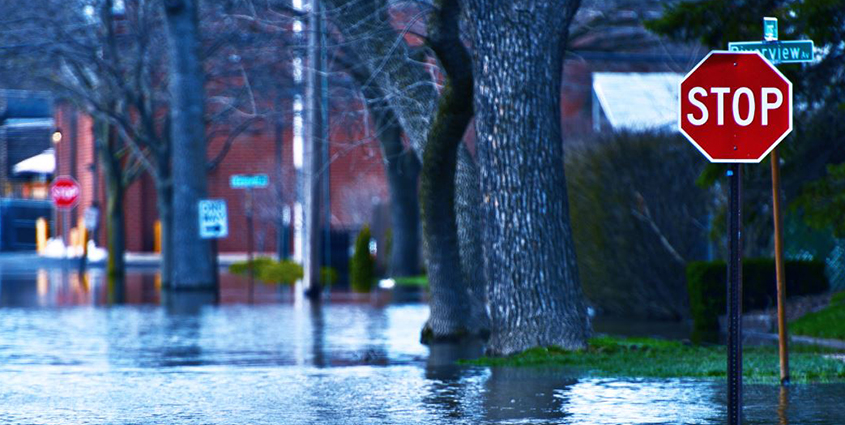 Deep water in flooded streets covering bottom of a stop sign at front of photo, large trees 