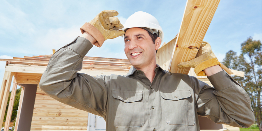 Smiling contractor tips white construction hard hat while carrying 2x4 